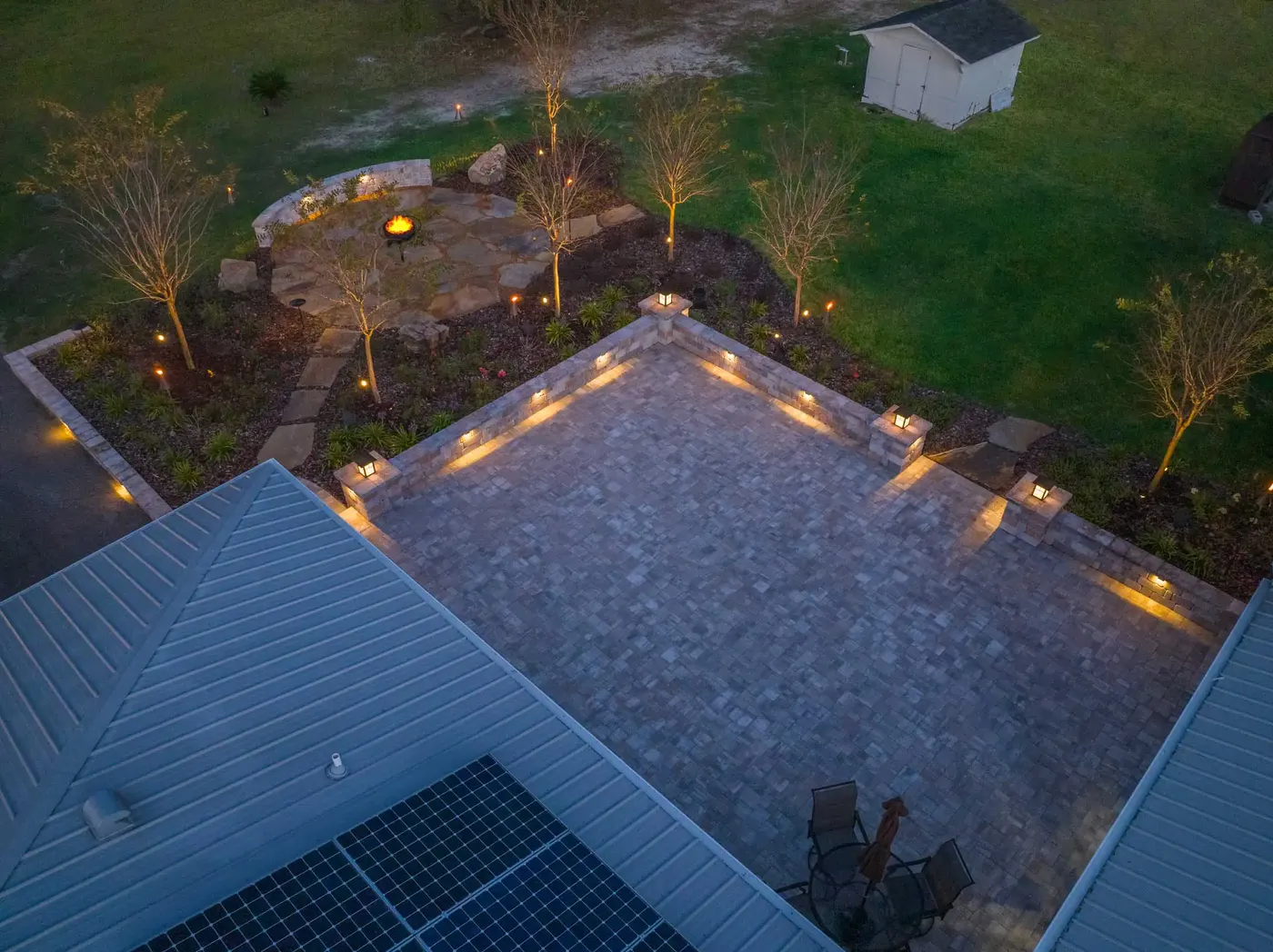 Drone view at night of a paver patio and beds lit by landscape lighting