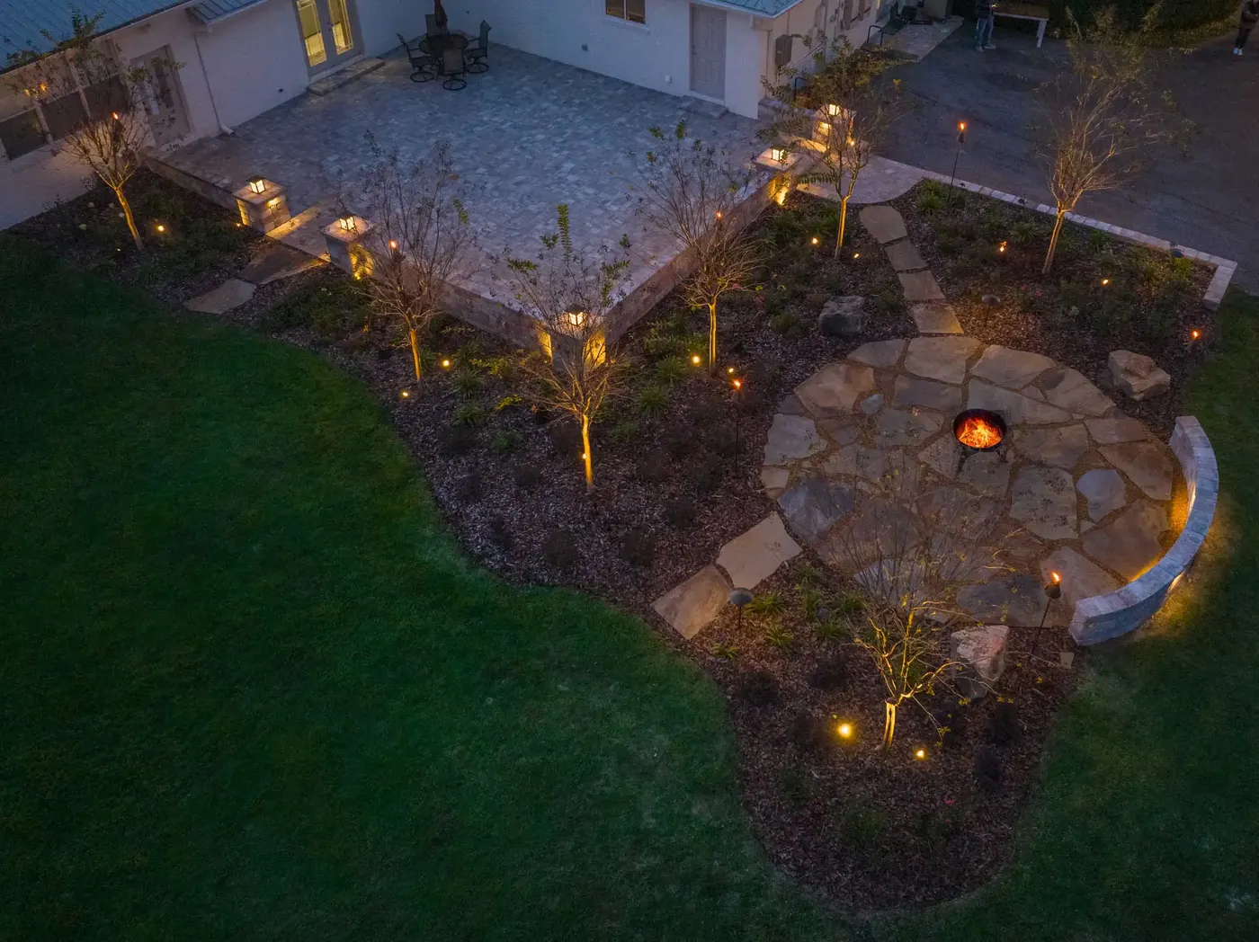 Night drone view of a lit flagstone fire pit patio and planted beds