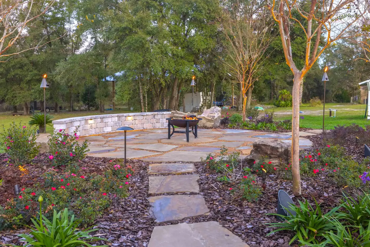 Paver patio with a fire table and stone seating wall under landscape lighting at dusk