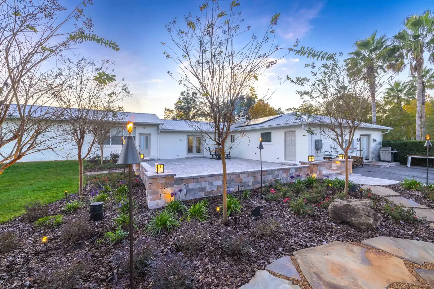 Paver patio and new plantings beside a white house at sunset, path lights on